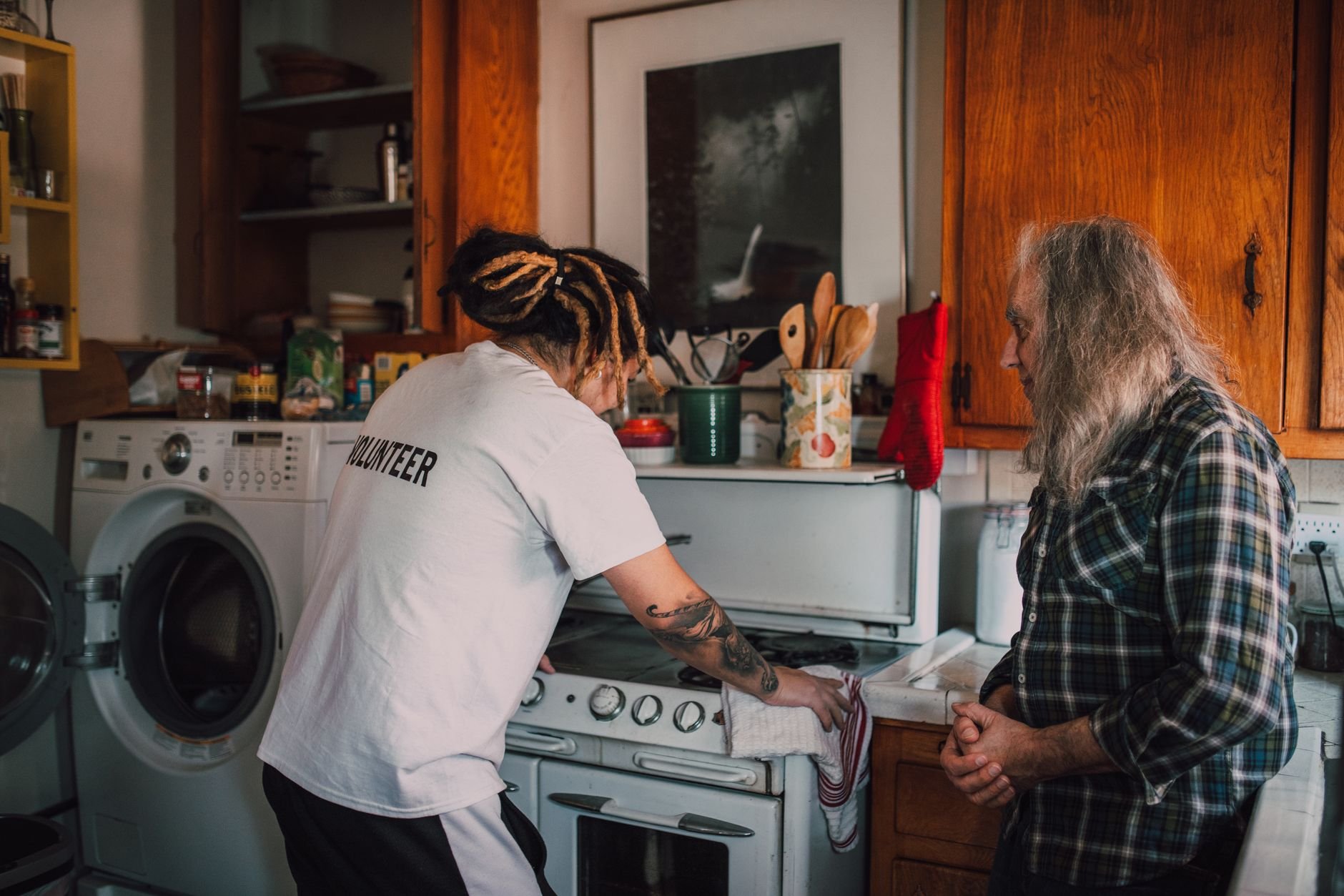 A volunteer helps an elderly person in a cozy kitchen, promoting community kindness.